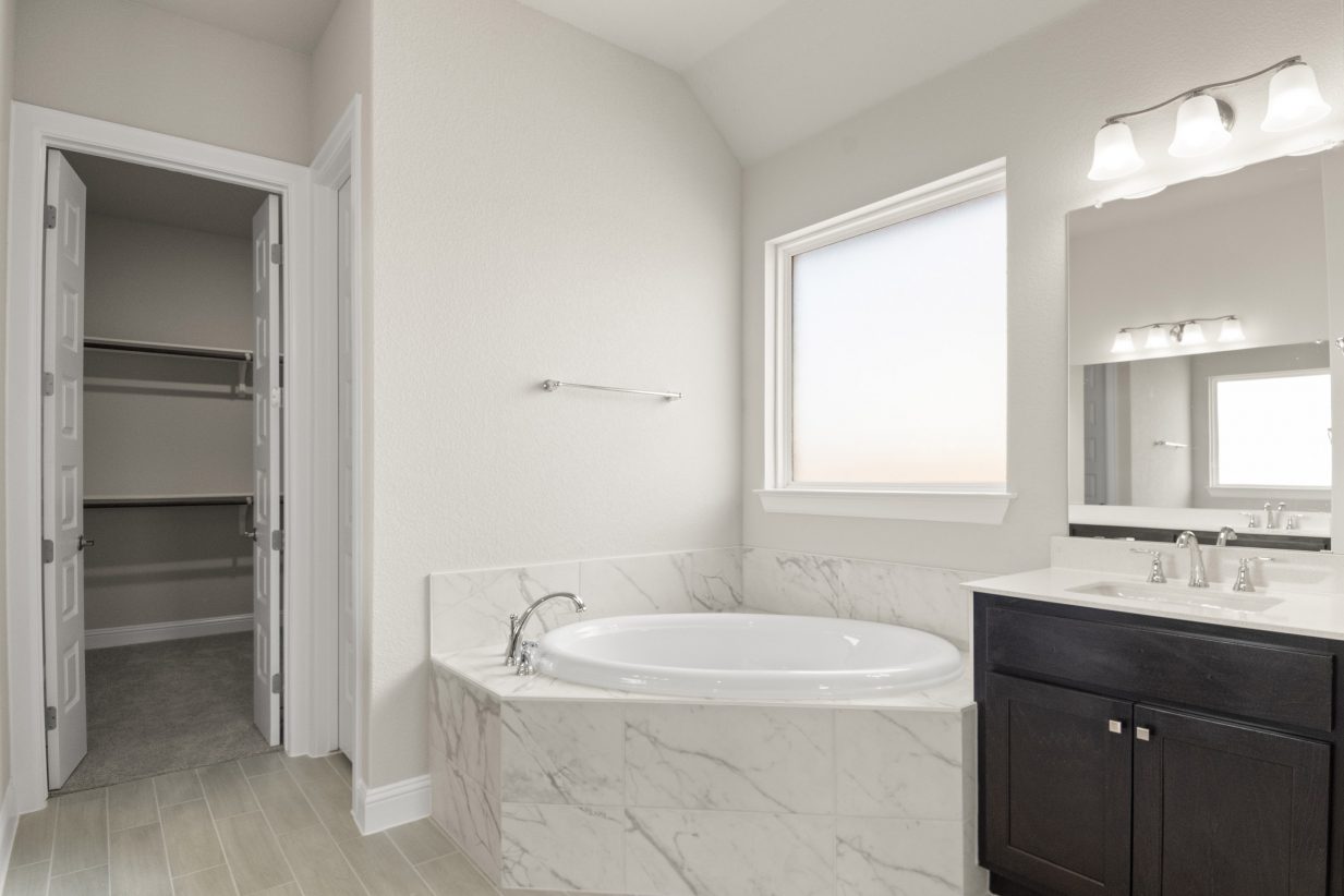 The primary bathroom with a marbled bathtub with a walk-in closet, a vanity with white countertop and dark brown cabinets, and vinyl flooring.