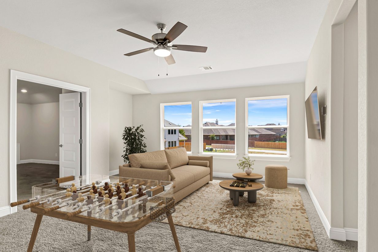 Image of a staged game room with a tan couch and rug with plant decor, a TV on the wall and a foosball table. The walls are painted grey with three windows and the floors are light grey and carpeted