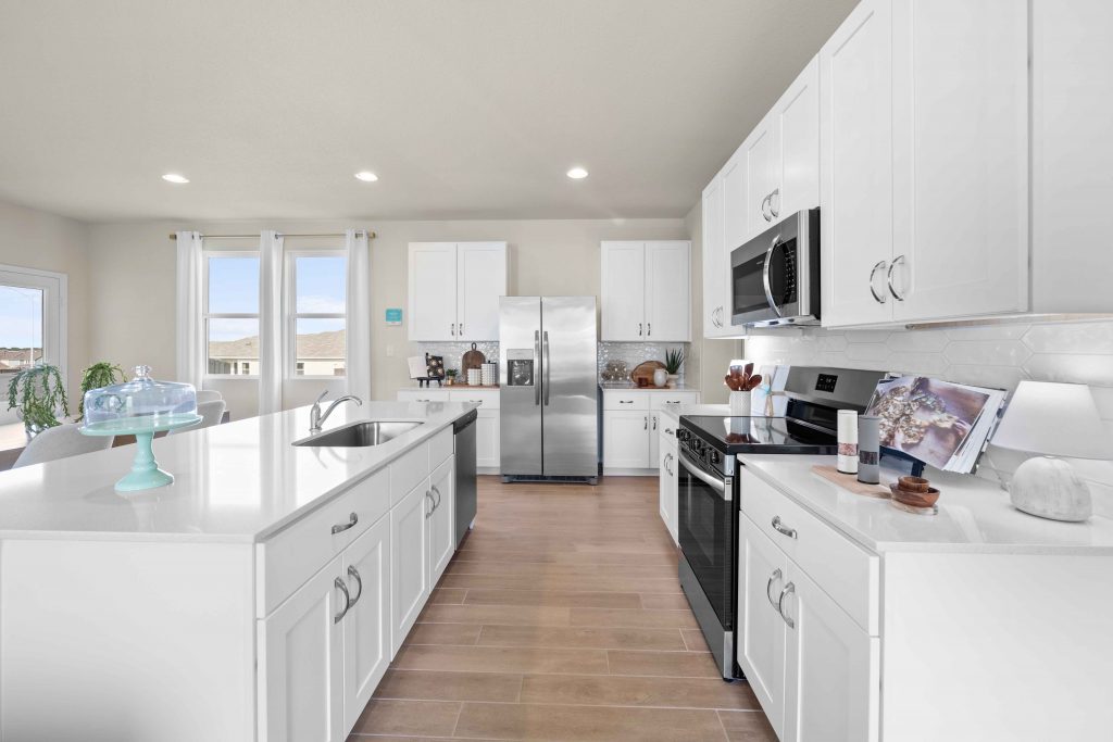 An interior image of a kitchen with white marble counter top and island, vinyl flooring, white cabinets, and steel refrigerator and dishwashers and a black and steel stove.