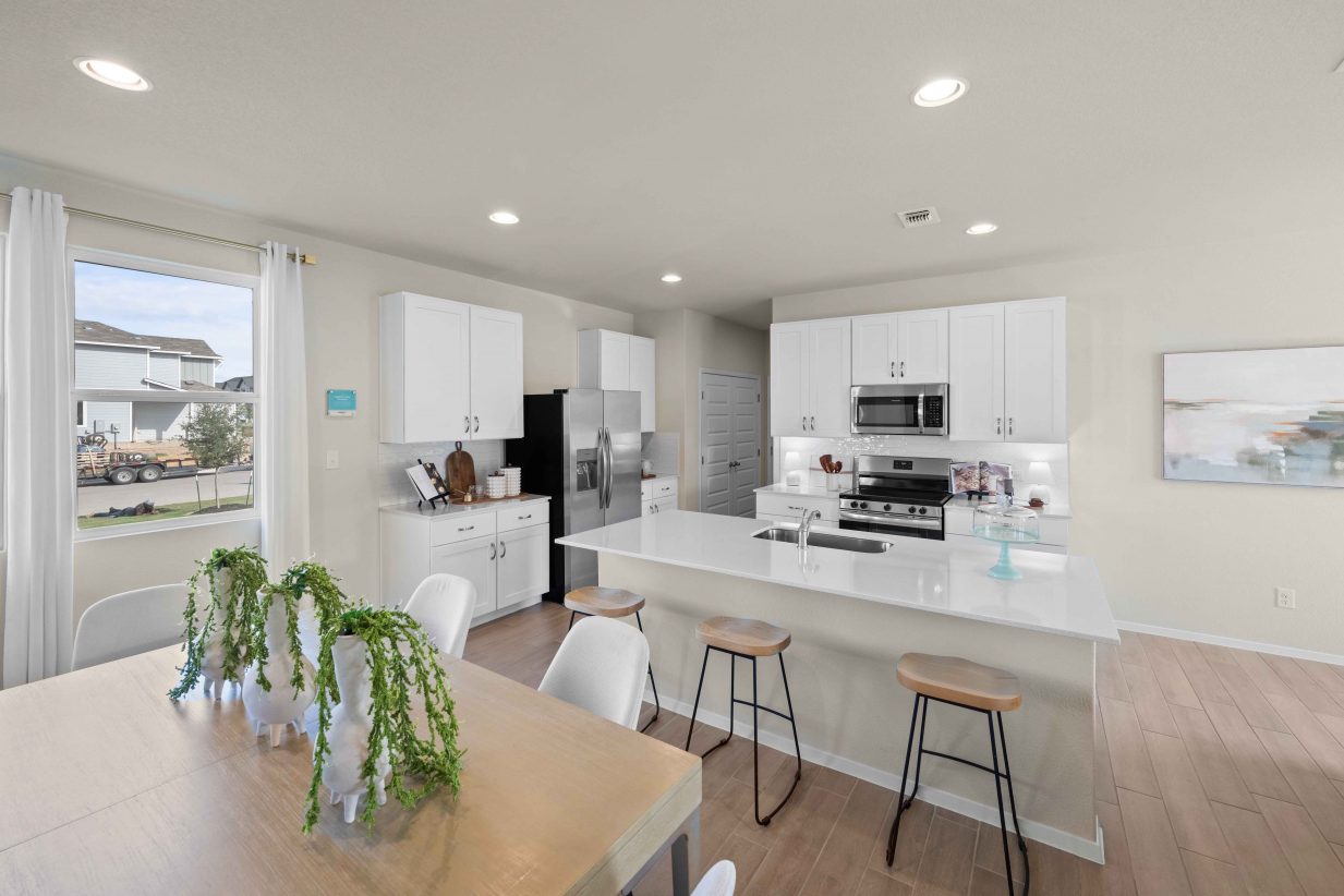An interior image of a tan dining table view of the kitchen with vinyl flooring, white cabinets and countertop.