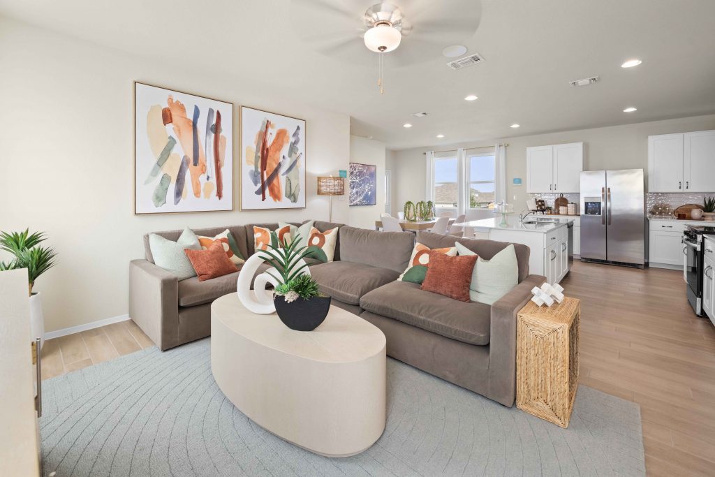 Livingroom with colorful pillows and art, a tan coffee table, grey run and green plants with a kitchen in the background.