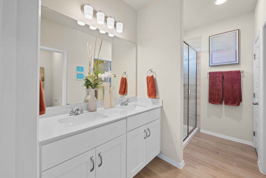 An interior image with a dual vanity with white countertops and white cabinets, a mirror, vinyl flooring, and glass shower door.