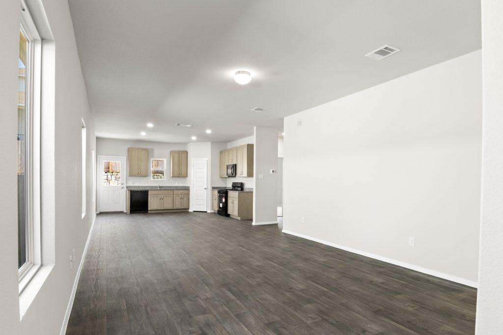 Image of a one story house living room with dark wood-like flooring and light grey painted walls with a kitchen in the distance