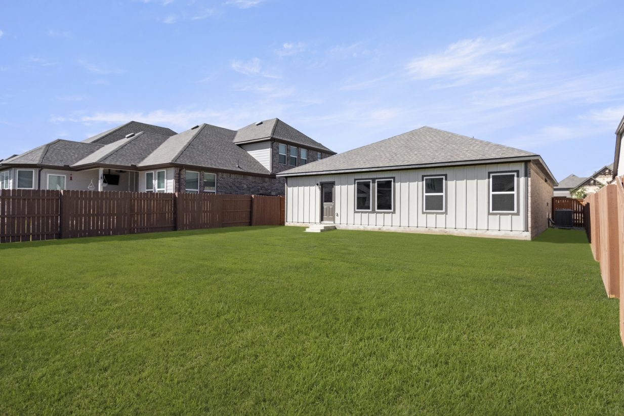 Image of the back exterior of a one-story home with a large green grass backyard, a brown wooden fence, and a blue sky in the background