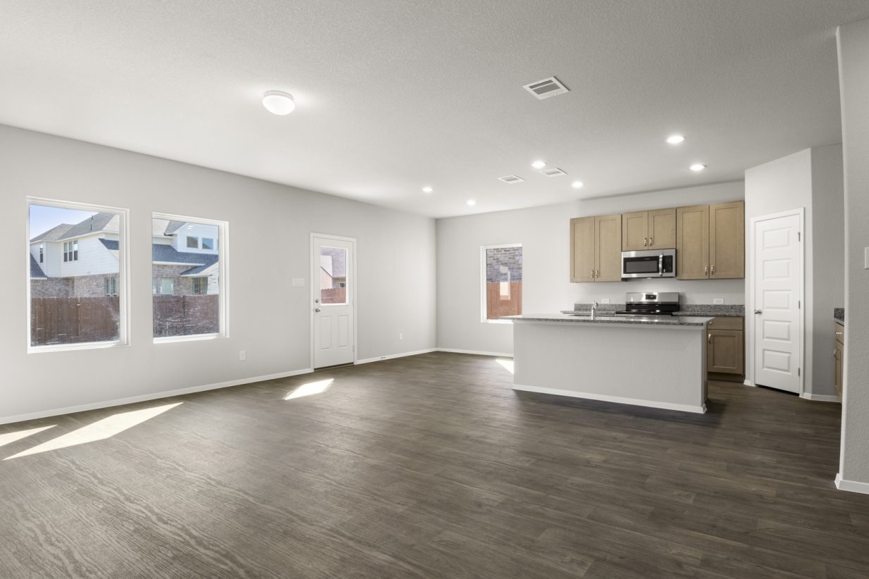 Image of a overview of a living, dining and kitchen area with dark wood-look flooring, light grey walls, windows and a back door