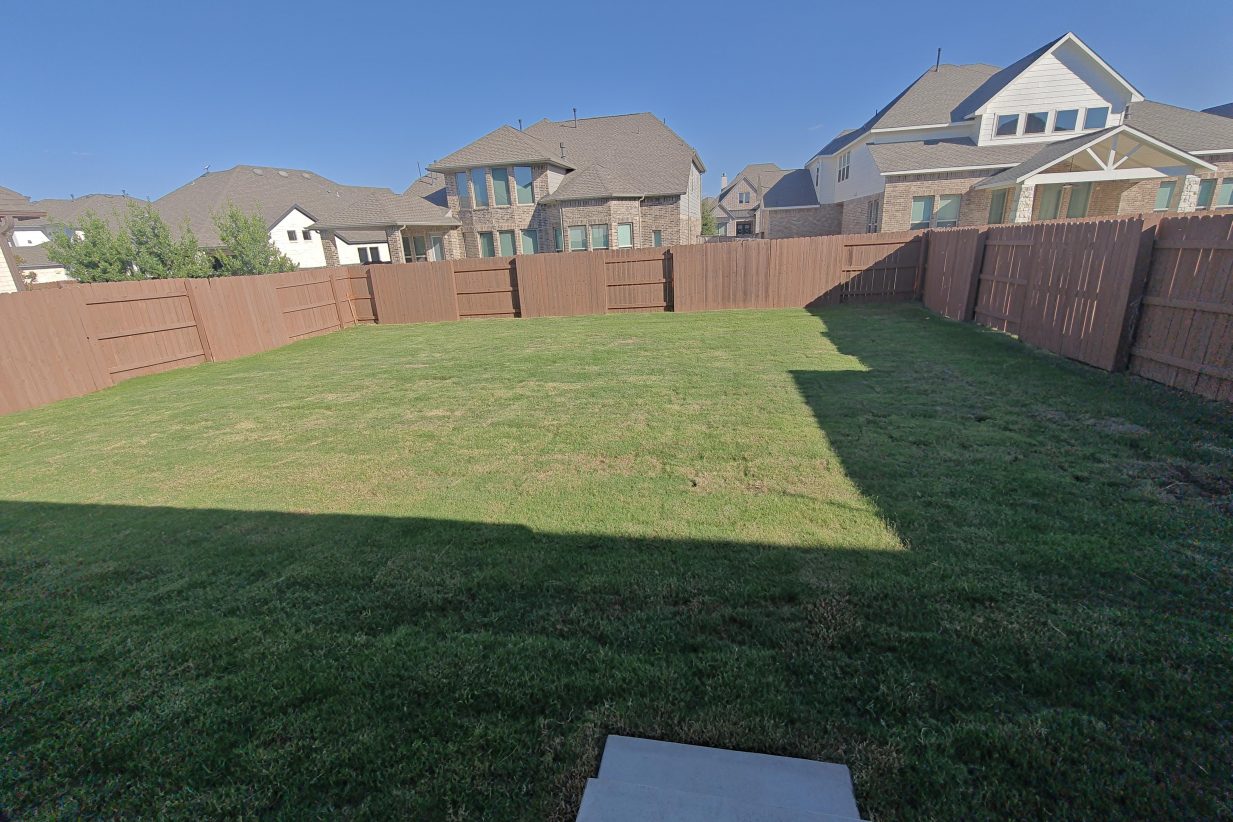 Image of backyard with green grass, a brown fence, houses in background with a blue sky.