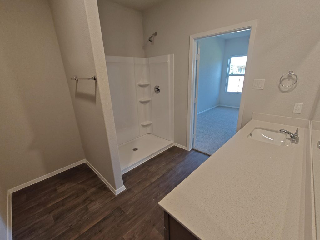 Image of a bathroom with brown vinyl flooring, brown cabinets with white countertops, painted white walls, and a white ceramic shower.