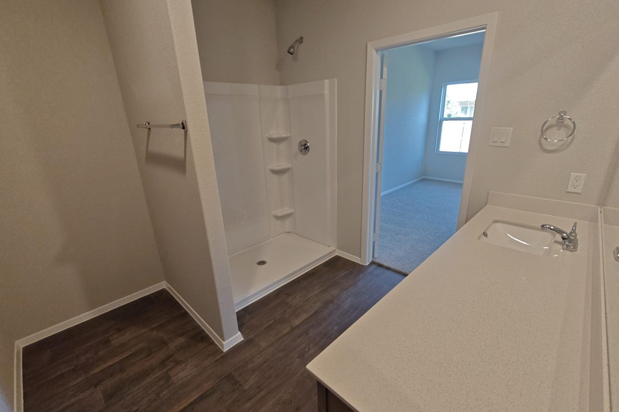 Image of a bathroom with brown vinyl flooring, brown cabinets with white countertops, painted white walls, and a white ceramic shower.