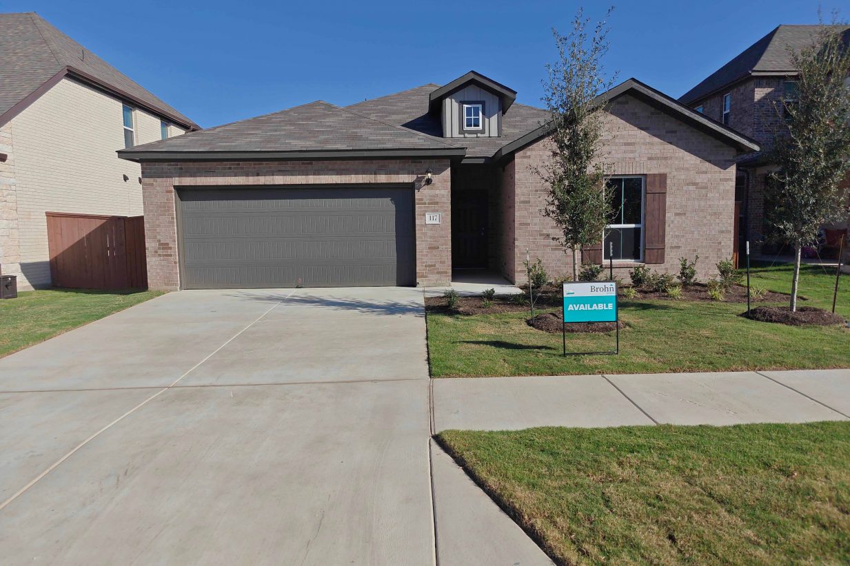 Image of front of a single-story brick house with a brown garage door, concrete driveway an d walkway, grass landscape, a tree, and teal "for sale" sign.