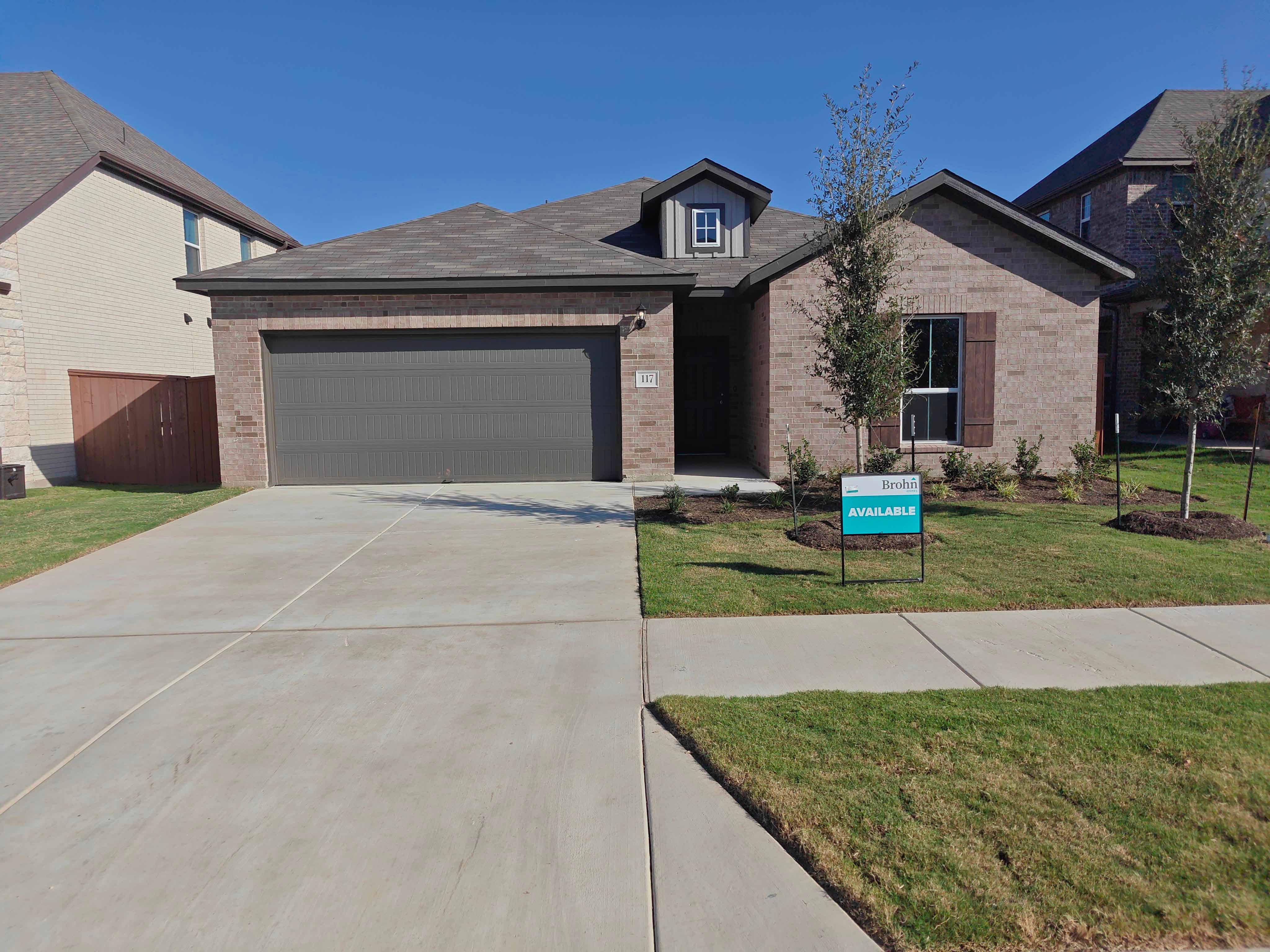 Image of front of a single-story brick house with a brown garage door, concrete driveway an d walkway, grass landscape, a tree, and teal "for sale" sign.