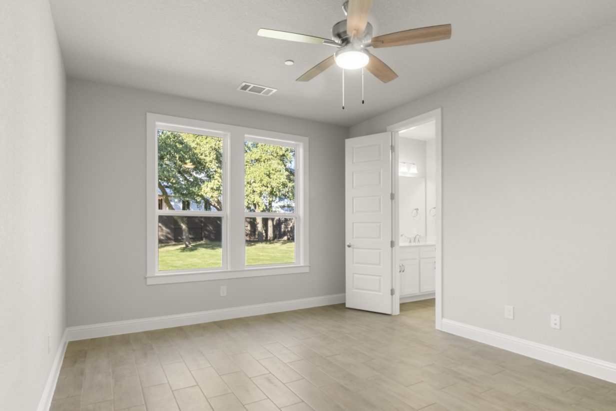 Image of primary bedroom with light grey painted walls, light brown wood-like flooring, two large windows, and a connecting bathroom