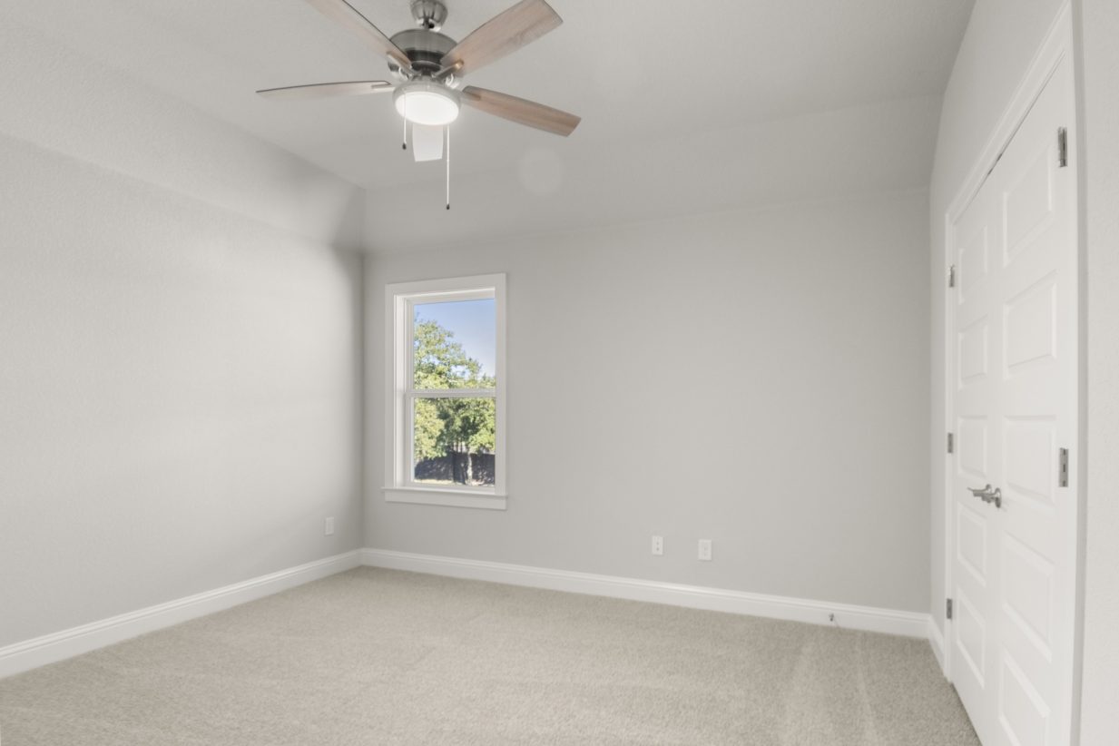 Image of a bedroom with tan carpeting and light grey walls with a window and a ceiling fan