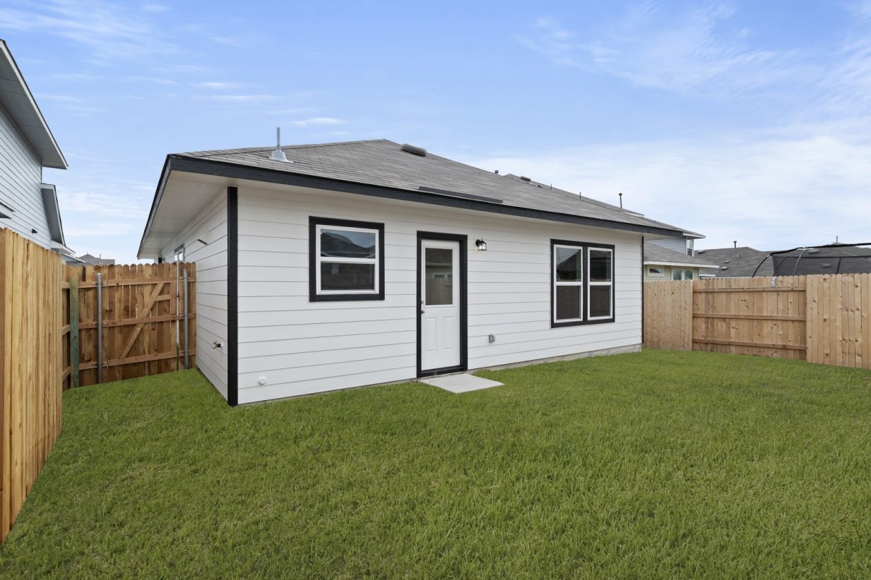 Image of a white one story house back exterior with black trim, windows, a green grass backyard, a wooden fence and a blue sky in the background