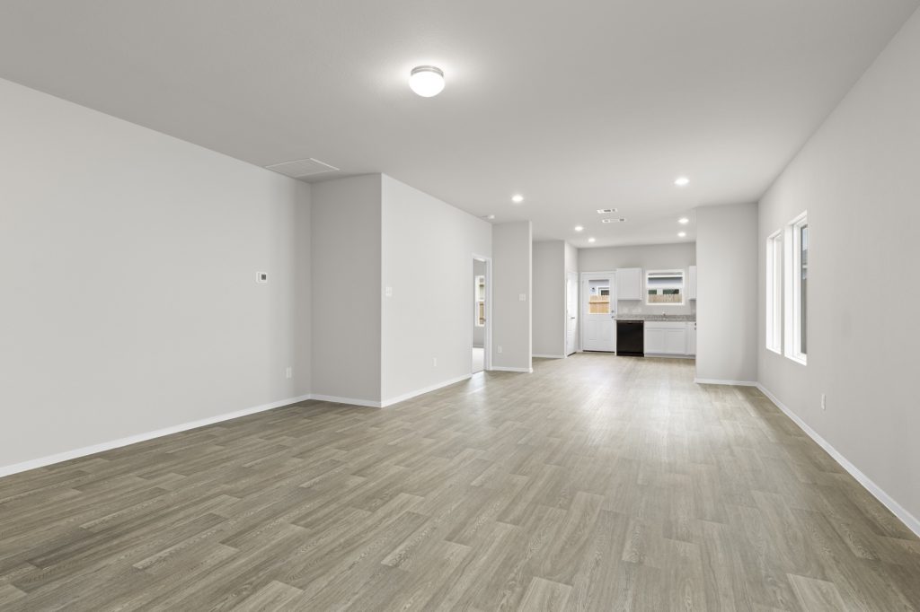 Image of a living room with beige walls, light brown vinyl flooring, and an open concept kitchen in the distance