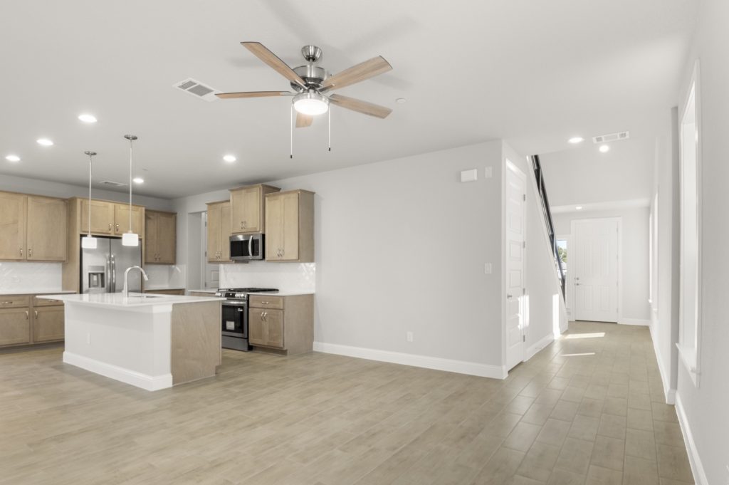 Image of a two story home living room with light brown flooring and grey painted walls with a front entrance walk way and staircase