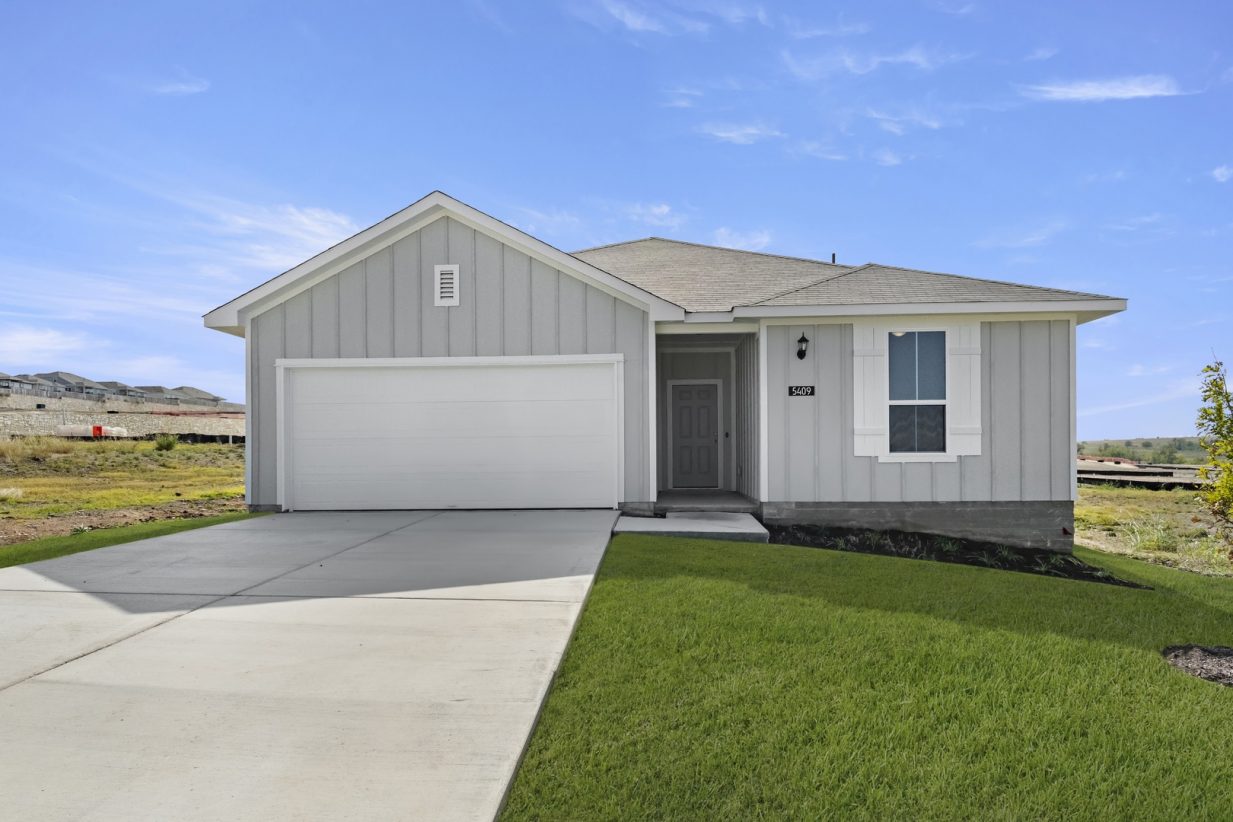 Image of the front exterior of a one story grey home with a white garage door, white shutters, a cement driveway and green grass with a blue sky in the background
