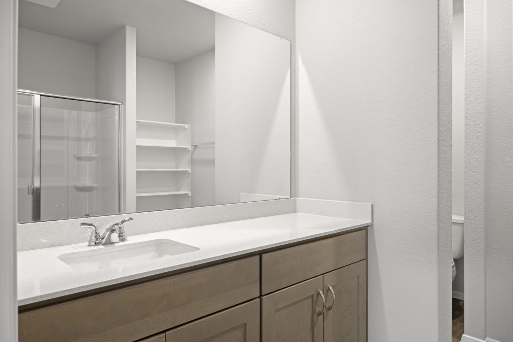 A bathroom with a white countertop, mirror, and brown cabinets, and white painted walls.