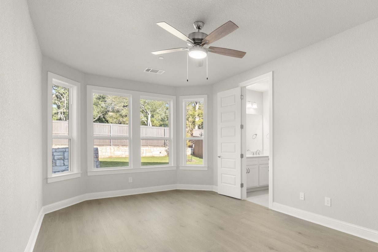 Image of a primary bedroom with light wood-like flooring and light grey painted walls with four large windows and a ceiling fan