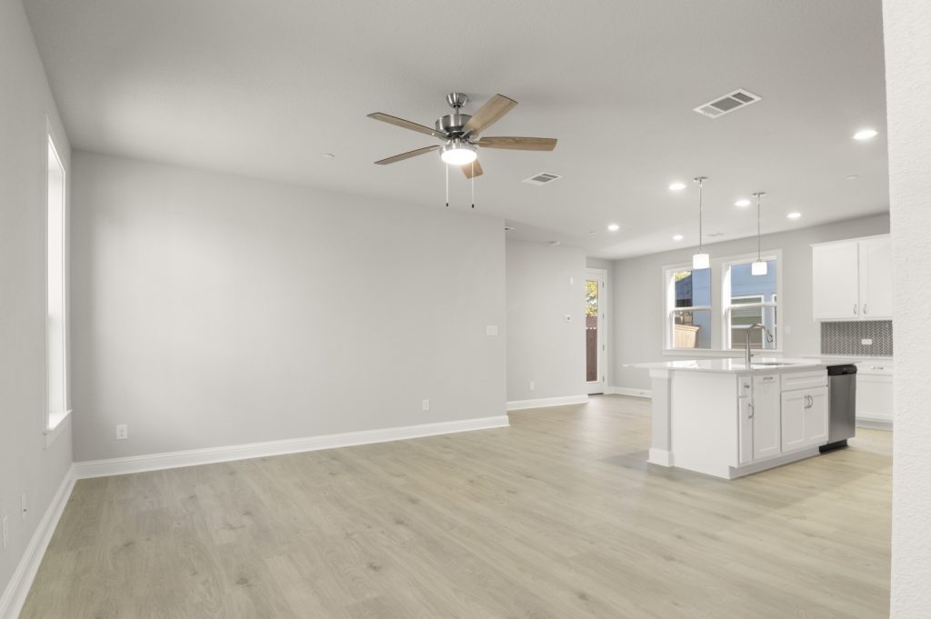 Image of a two story home living room with light wood-like flooring and light grey painted walls with a kitchen connecting