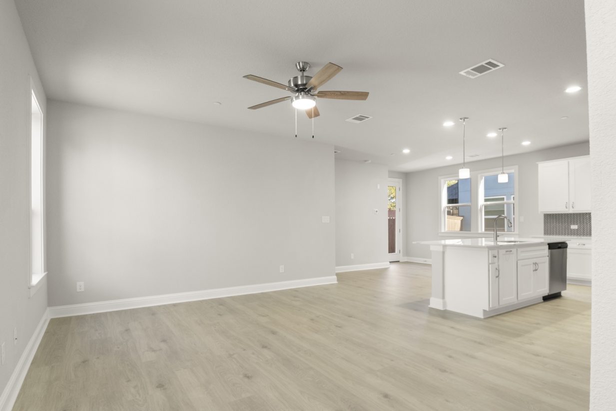 Image of a two story home living room with light wood-like flooring and light grey painted walls with a kitchen connecting