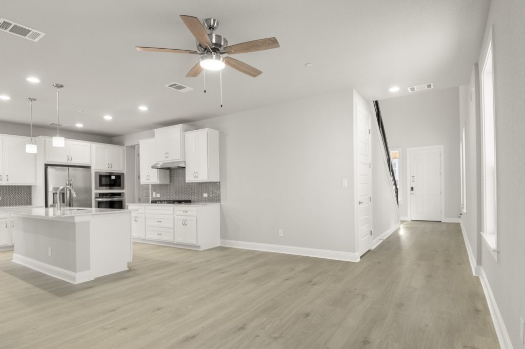 Image of a two story home living room with light wood-like flooring and light grey painted walls with a kitchen connecting