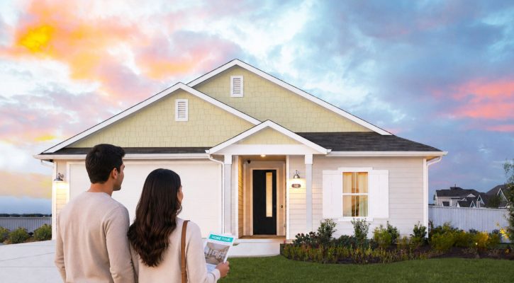 A picture of the back of couple wearing tan sweaters with brunette facing a one story yellow home with white trimming and a white garage door with a colorful sky with clouds in the background.