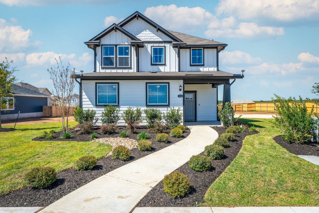 An exterior image of white two-story home with black framing, windows, with a concrete walkway with a green grass landscape surrounding and a blue sky with clouds.