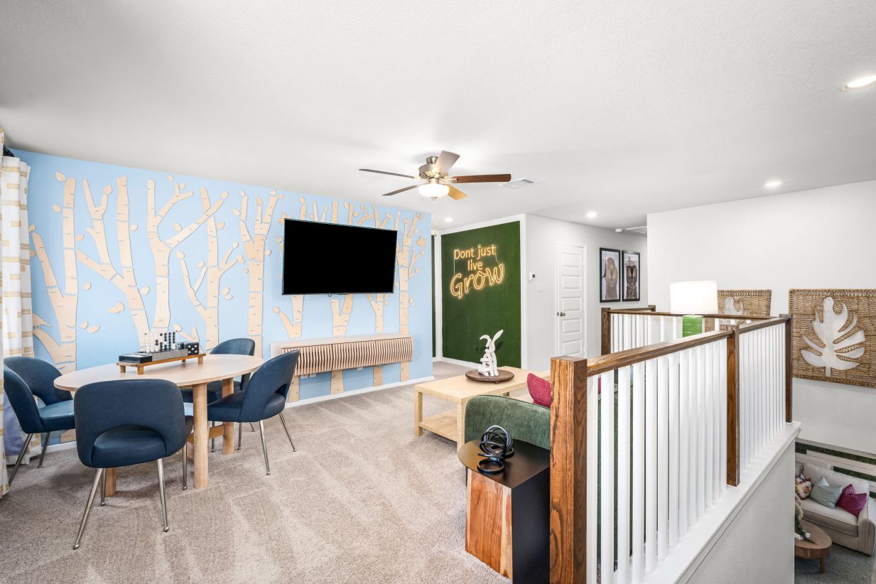 Image of a recreation room with a light blue wall with tree decoration, tan carpet, an accented wall with greenery, brown ceiling fan, with a table with black chairs, green couch with colorful pillows.