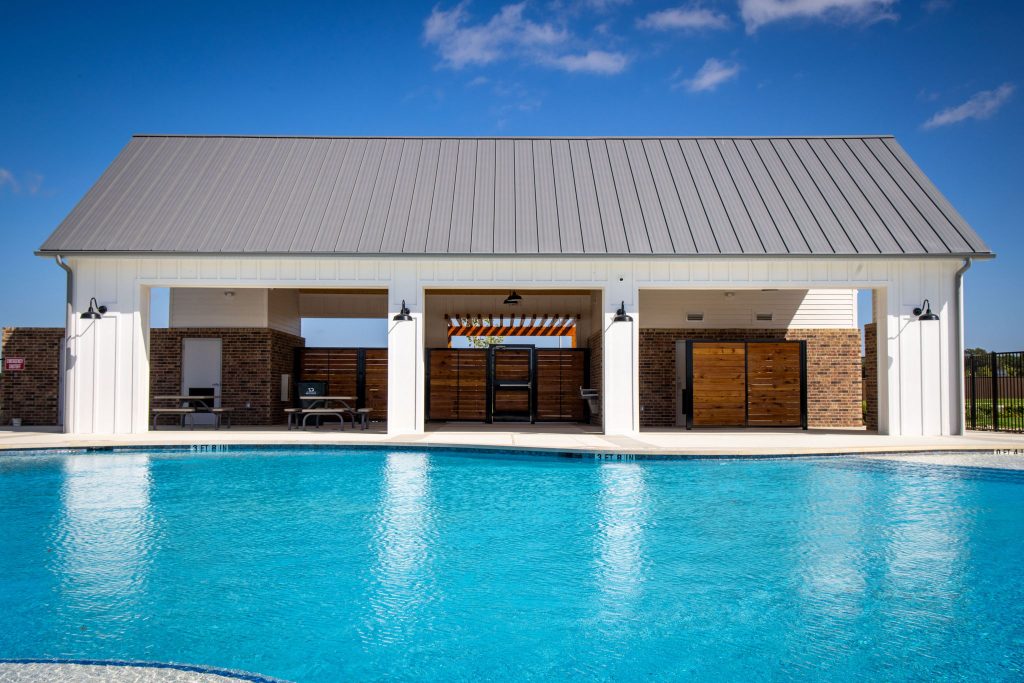An image of a pool with blue water and a concrete pool deck, a white and gray building, and a blue sky with clouds.