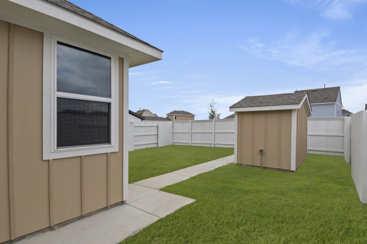 Image of a back exterior of a tan one story home with a small shed, green grass and a white fence