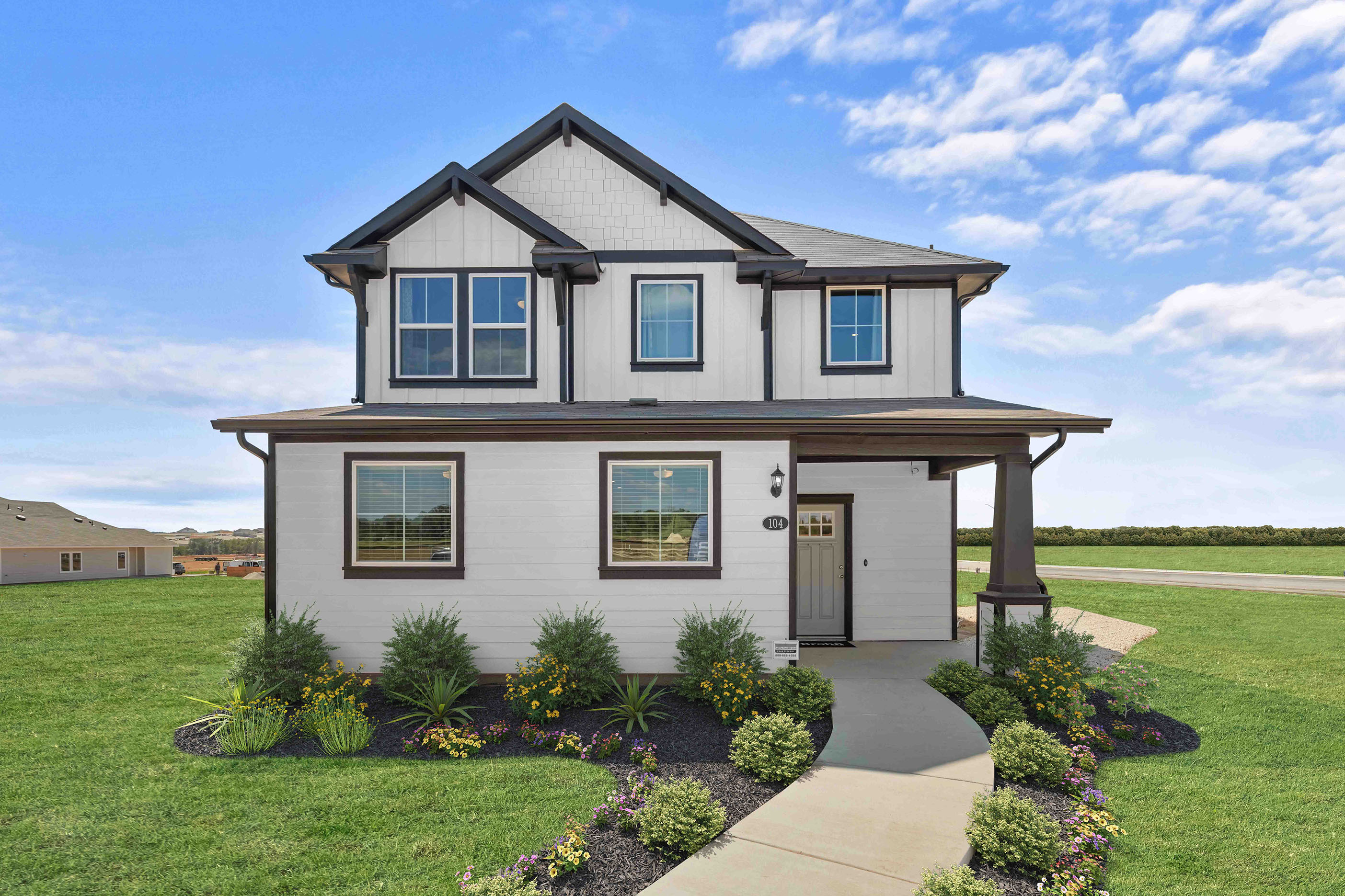 Image of front exterior of two-story house with grass landscape, bushes, flowers, blue sky with clouds, and concrete sidewalk.