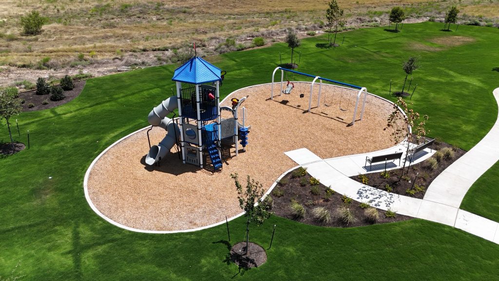 Aerial photo of blue and grey community playground including a slide and swings surrounded by playground mulch with a sidewalk surrounded by grass leading to it