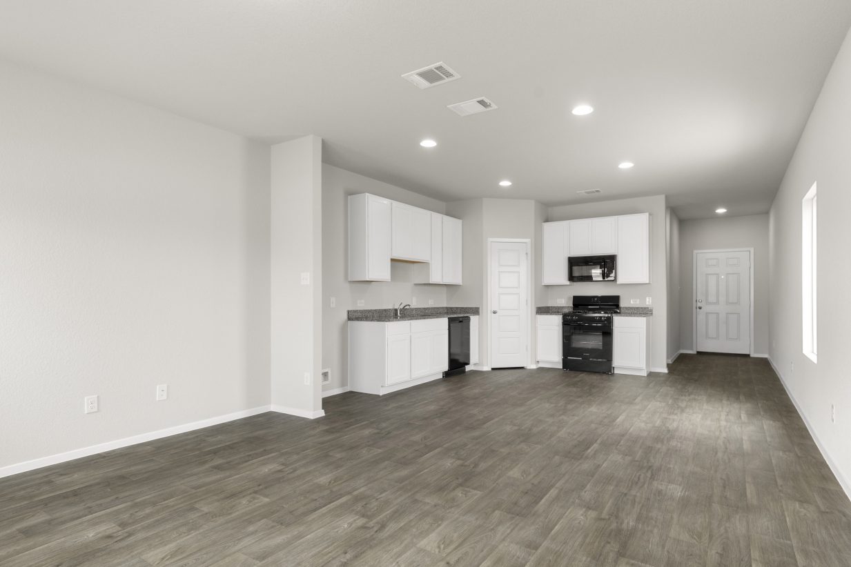 An image of a kitchen and living room with wood vinyl flooring, cream colored walls, white cabinets with grey countertops, and black appliances.