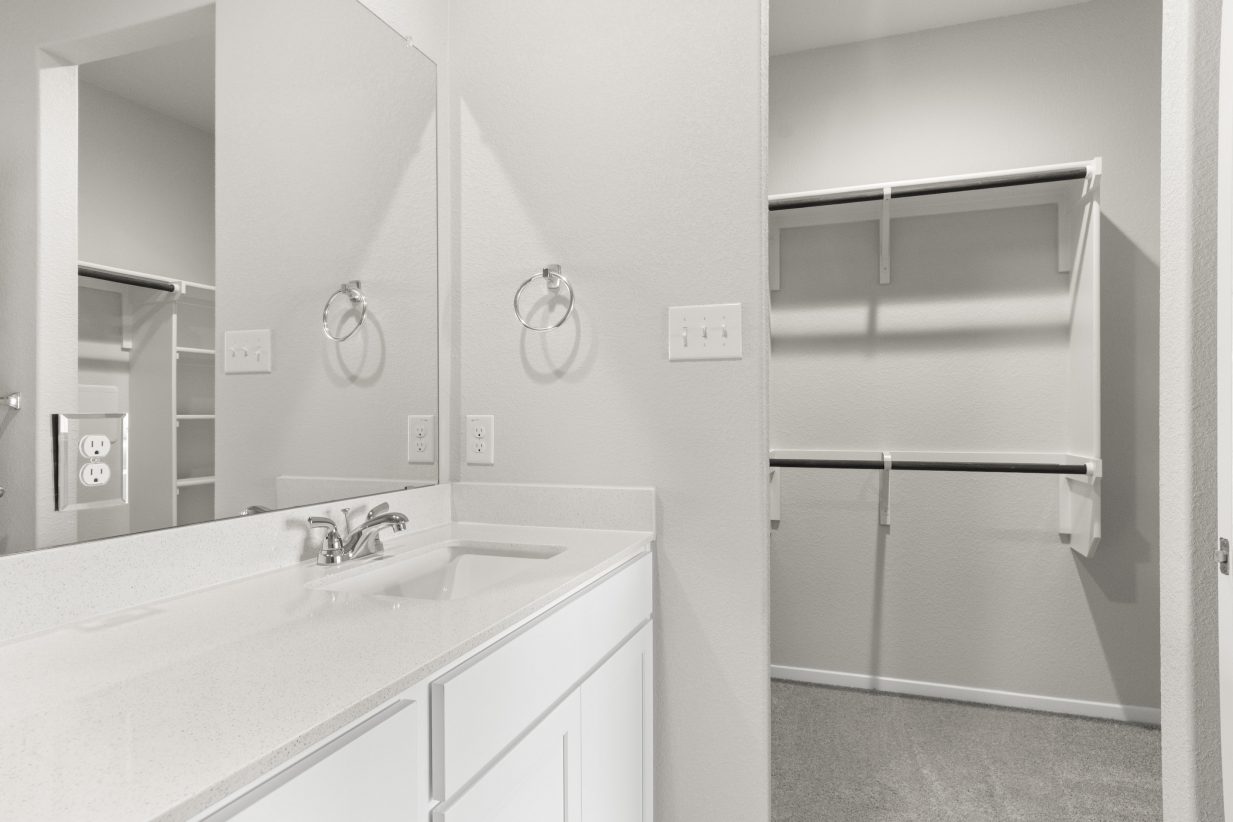 An image of a bathroom with a white vanity with white countertops, a mirror, a door way leading to a walk-in closet, and cream colored painted walls.
