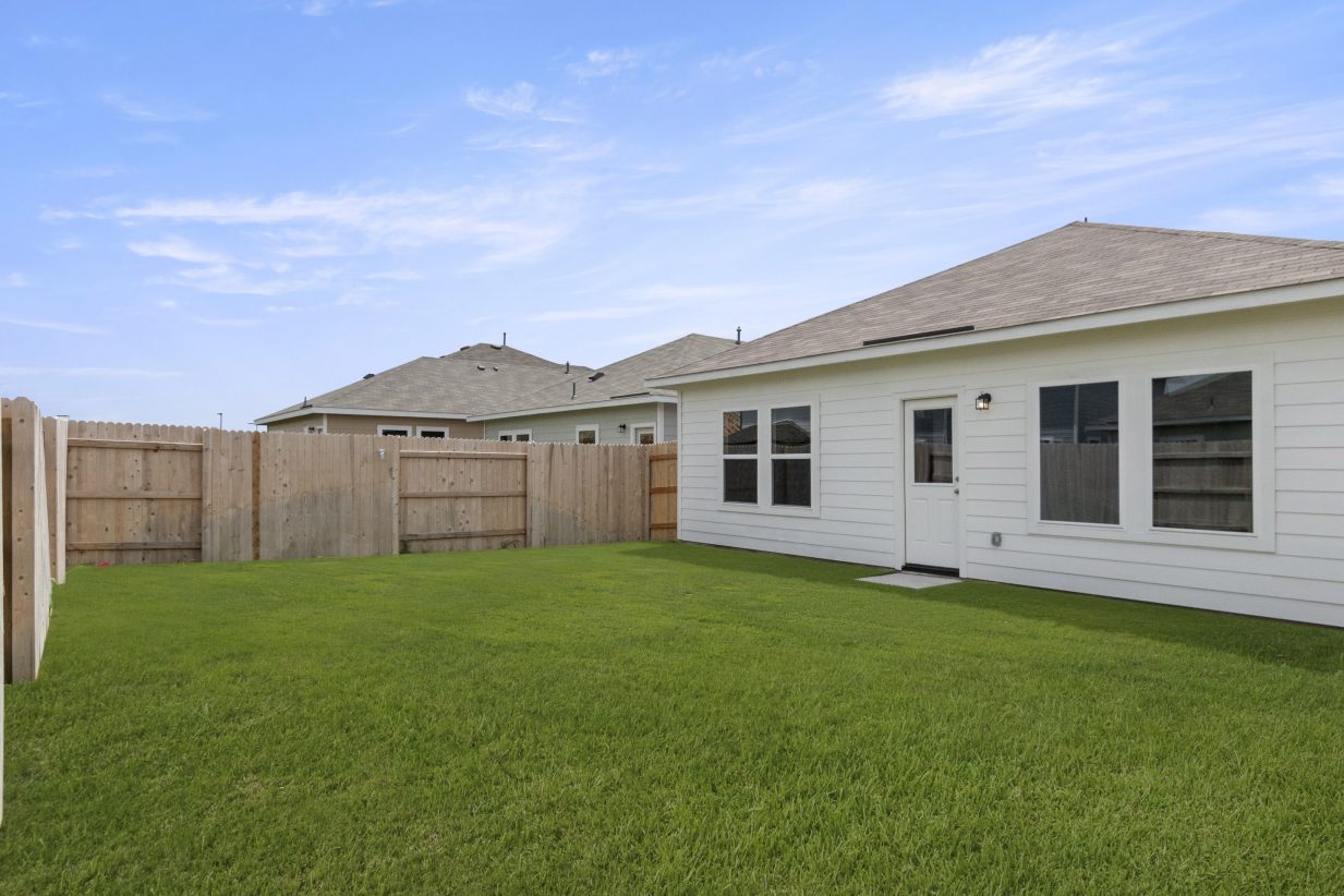 An image of a backyard with green grass, a light brown fence, the back of a white house, and a blue sky background.