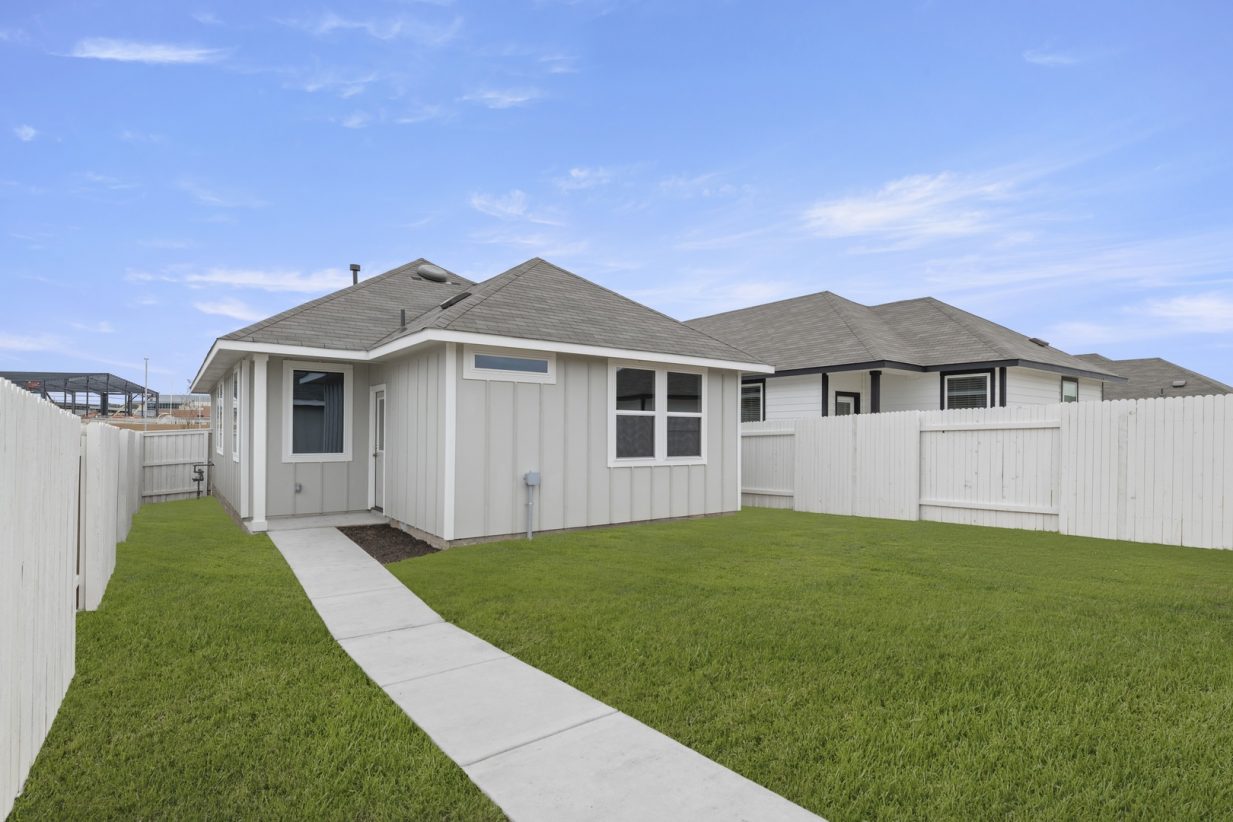 Image of a light grey one story house back exterior with a green grass backyard, a white wooden fence, and a blue sky in the background
