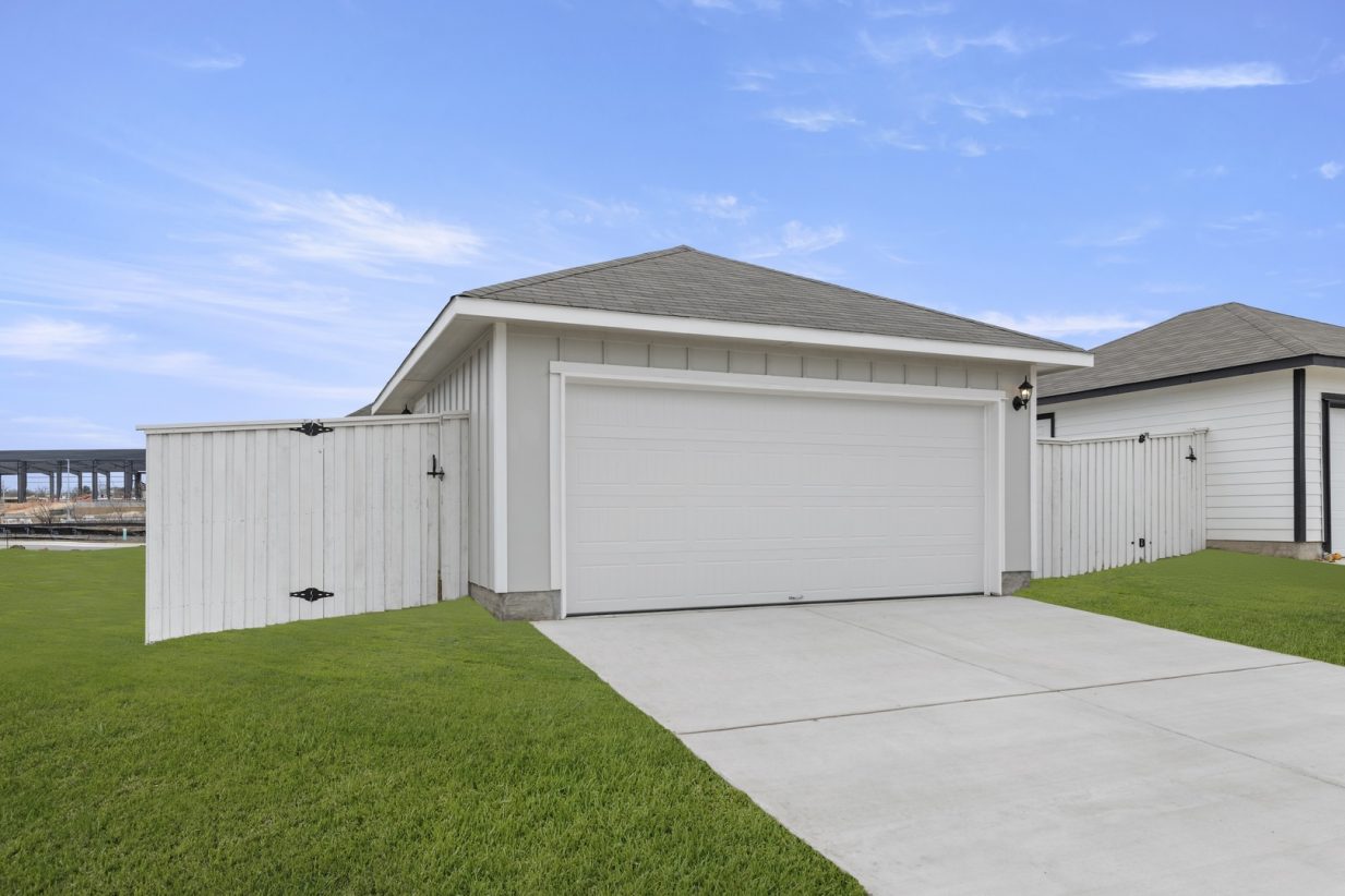 Image of a light grey garage with a white door, a cement driveway, a white wooden fence and a blue sky in the background