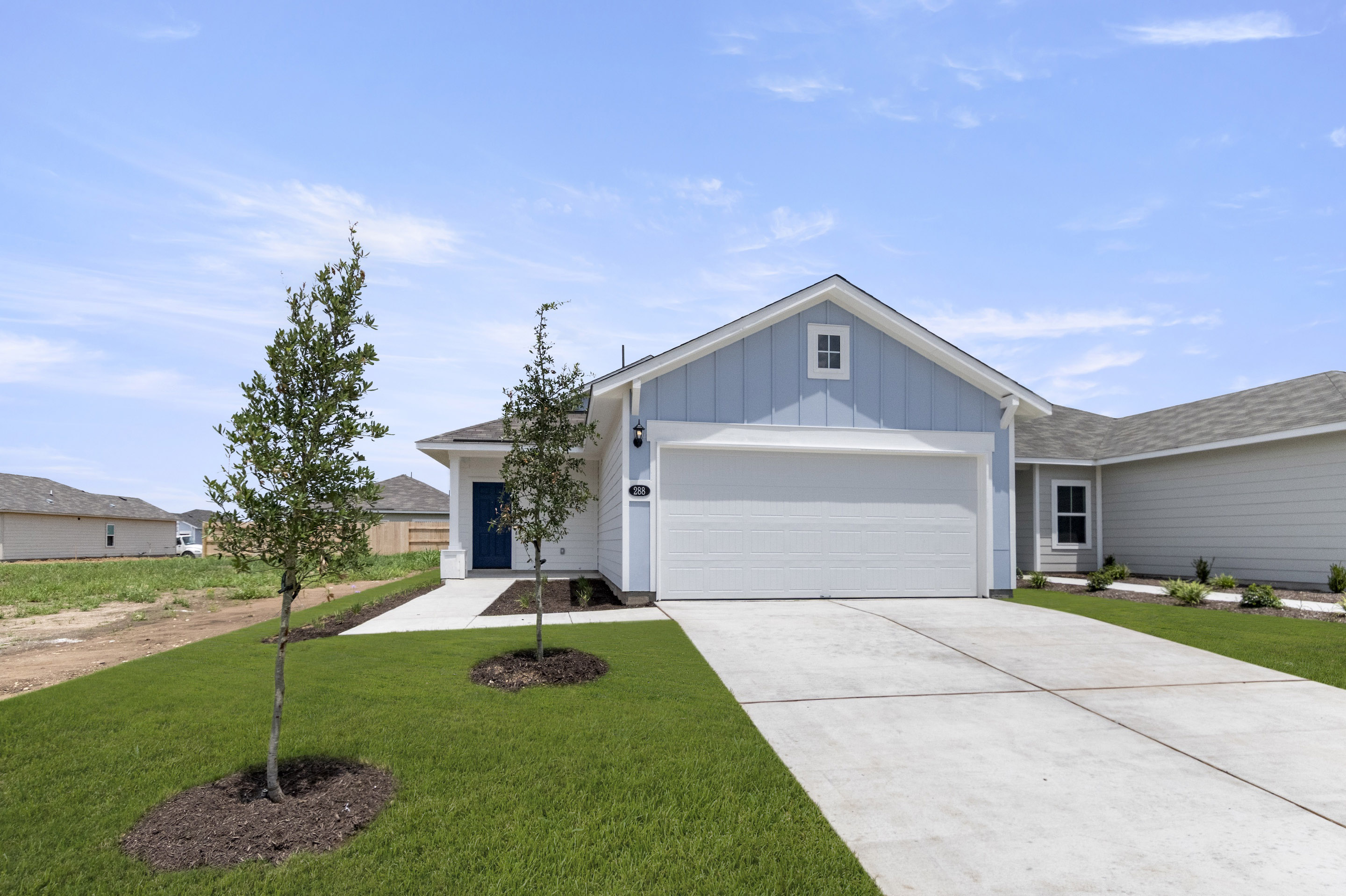 An image of the front of a blue single-story house with a white garage doors, dark blue front door, grass landscape, trees, concrete driveway, and blue sky background.