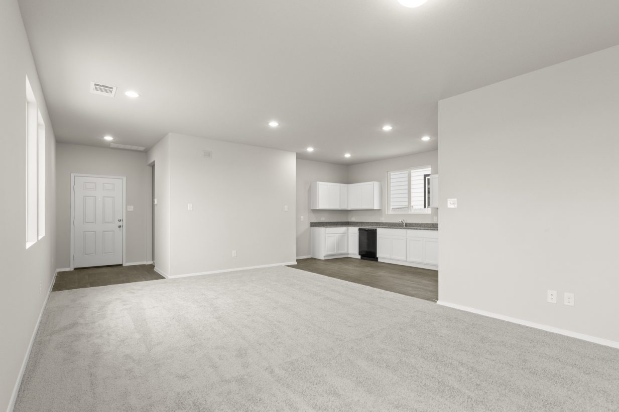 Image of a living room with cream walls, light grey carpeting, and a kitchen to the right with white cabinets