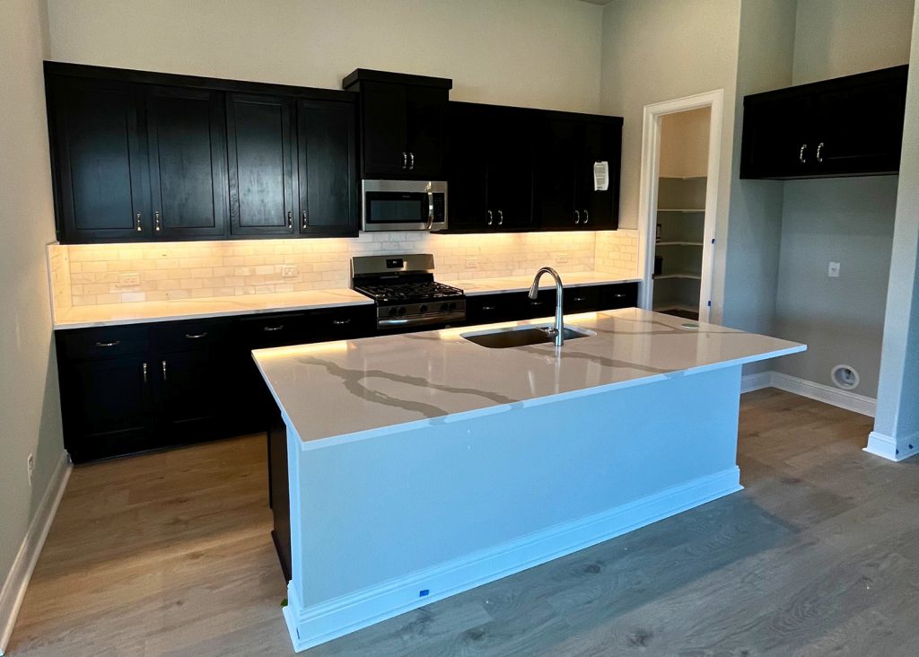 Image of kitchen with wood flooring, brown cabinetry, stainless steel kitchen appliances, white tiled blacksplash, a center island with white marbled countertop and a sink.