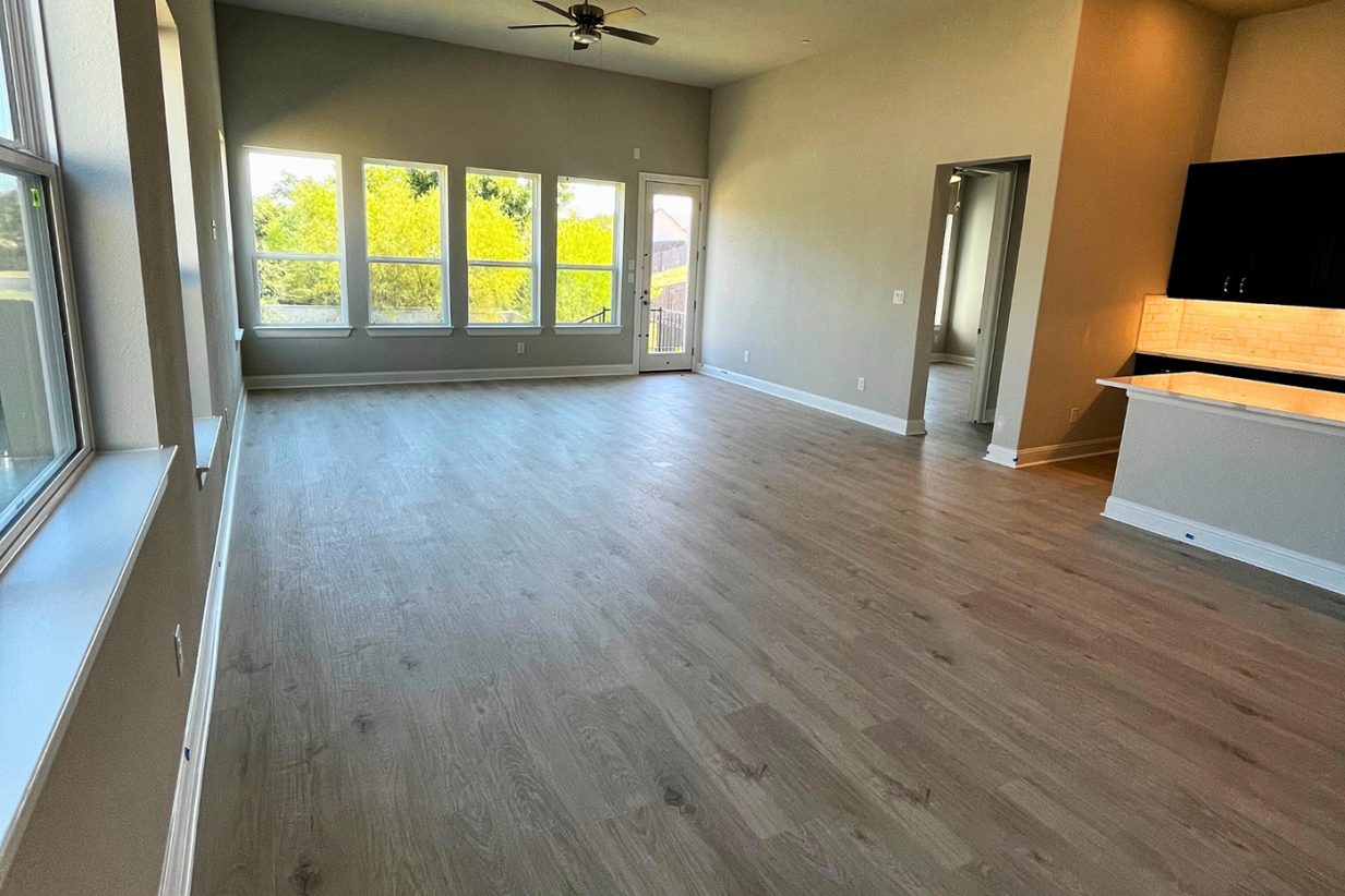 Image of living room and kitchen with wood flooring, with tan painted walls, brown ceiling fan, white framed windows, and a white countertop island.