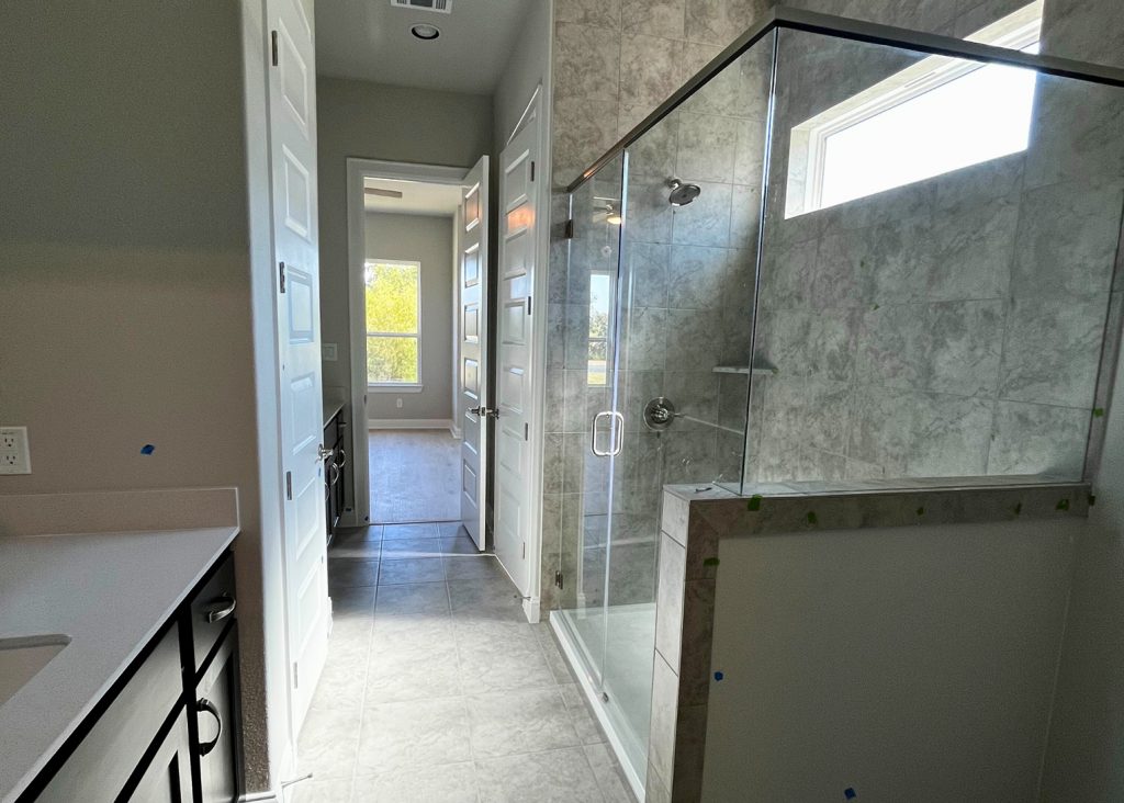 Image of a primary bathroom with marble tiled glass walk-in shower, gray marbled tiled flooring, white countertop vanity with brown cabinets, and tan painted walls.