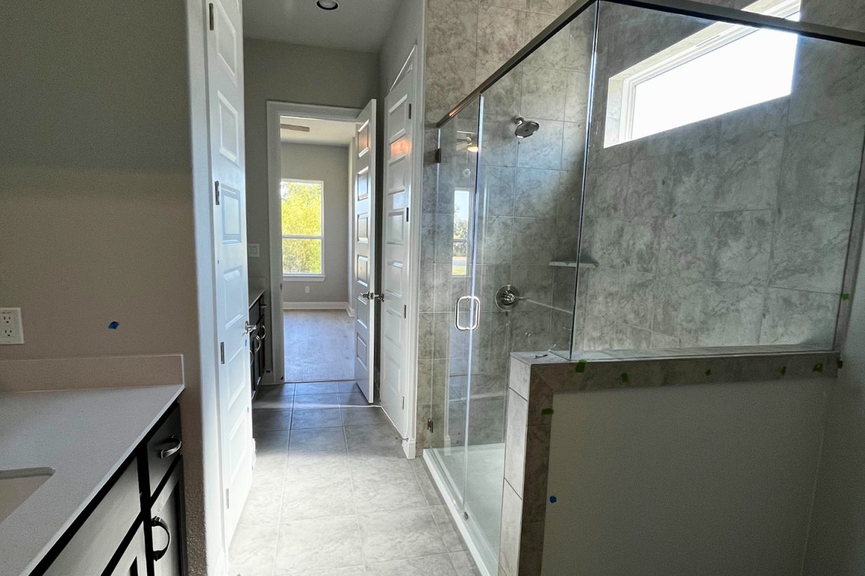 Image of a primary bathroom with marble tiled glass walk-in shower, gray marbled tiled flooring, white countertop vanity with brown cabinets, and tan painted walls.
