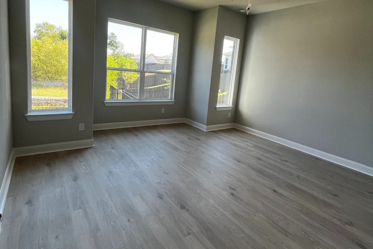 Image of a primary bedroom with wood flooring, tan painted wall with white base boards, and white framed windows.