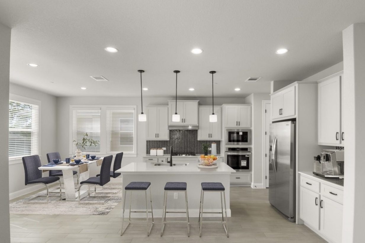 Image of kitchen and dining interior with a white modern dining room table with blue chairs and a dining set. Kitchen appliances and fruit on the counter