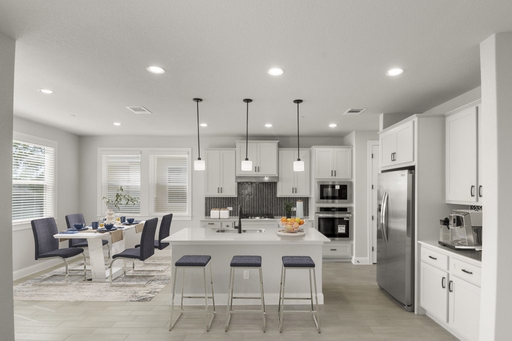 Image of kitchen and dining interior with a white modern dining room table with blue chairs and a dining set. Kitchen appliances and fruit on the counter