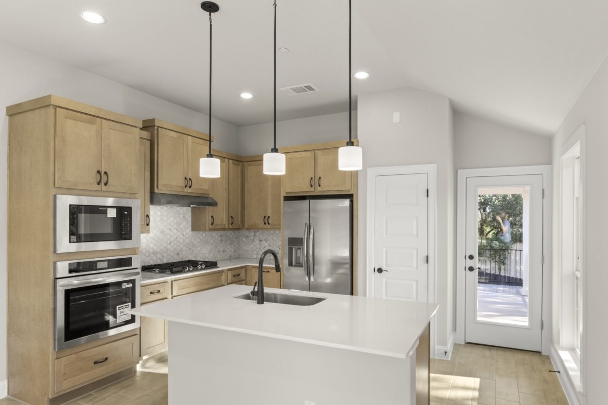 Image of a one story home kitchen with a white center island with a sink, light brown cabinets, marble backsplash, and stainless steel appliances