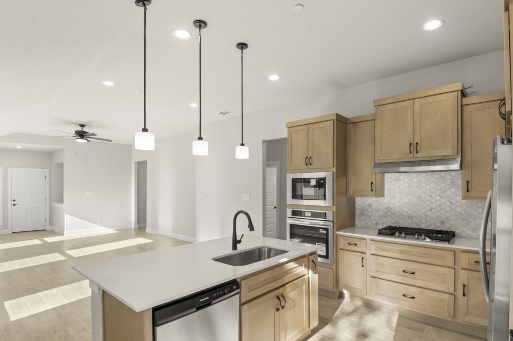 Image of a one story home kitchen with a white center island with a sink, light brown cabinets, marble backsplash, and stainless steel appliances