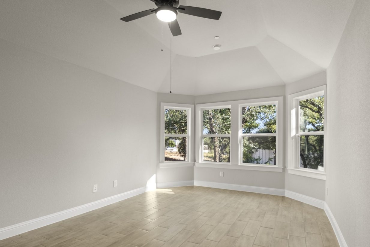 Image of a primary bedroom with light wood-like flooring and light grey painted walls, four large windows, and a black ceiling fan