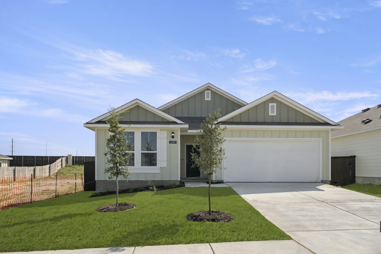 Image of one-story grey-green home with white trim and garage door, and black front door. Driveway, sidewalk, landscaping including two trees